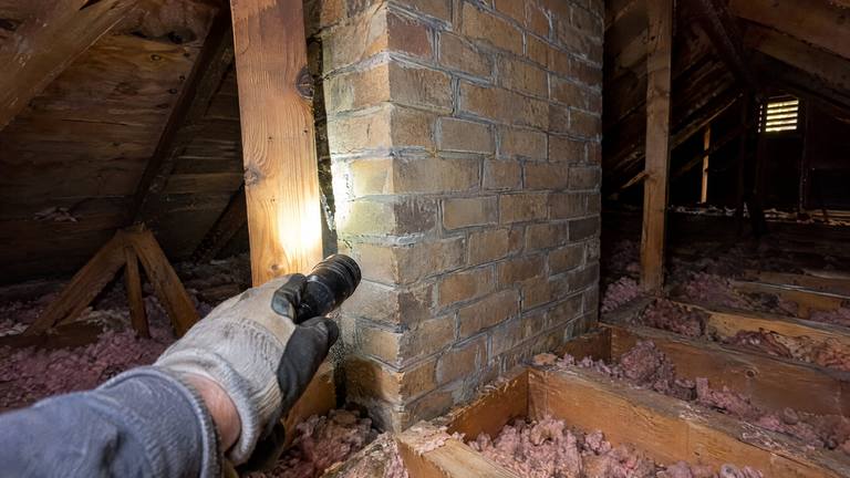 A flashlight in an attic lights the brick chimney where it passes between the roof rafters.
