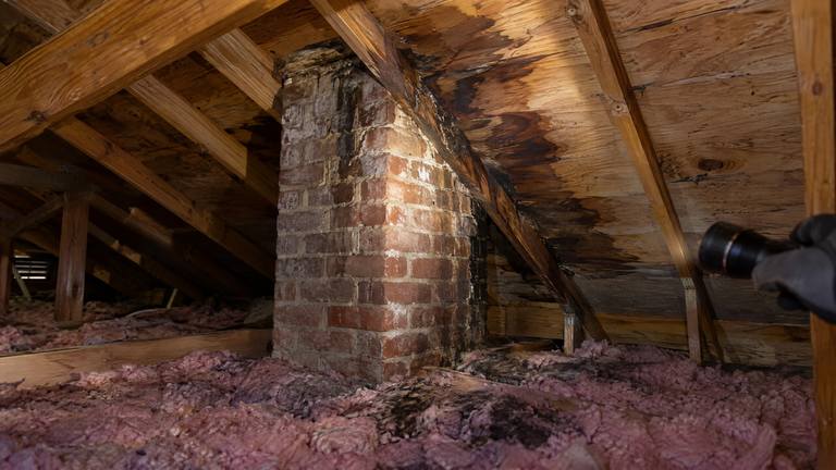 Dark water stains on the roof decking, rafters and insulation around a brick chimney inside an attic, lit by a flashlight.