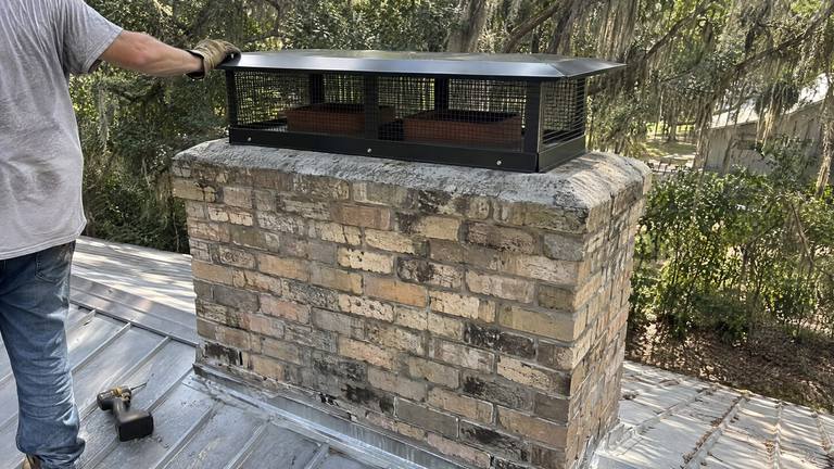A black multi-flue cap covers two flues on a wide tan brick chimney rising from a metal roof.