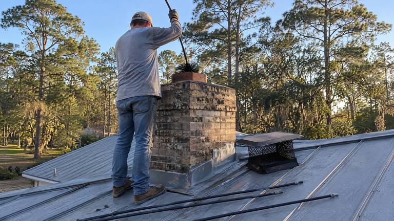 A worker on a metal roof pulls a round chimney brush on a rod out of the flue of a tan brick chimney.