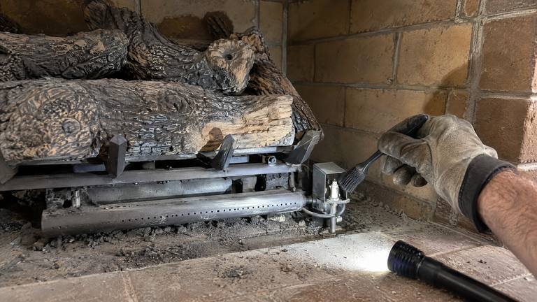 A gloved hand brushes dust from the small pilot of a vent-free gas log set.