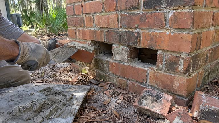 Gloved hands spread mortar on a new brick beside gaps at the base of an orange-red brick chimney.