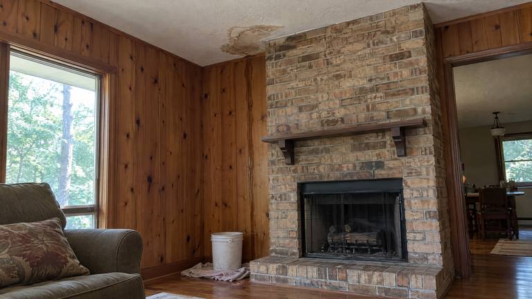 A brown water stain spreads across the ceiling beside a tan brick chimney breast in a paneled den.
