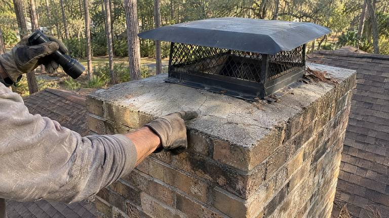 A gloved hand rests on the cracked concrete crown of a tan brick chimney while the other holds a flashlight.