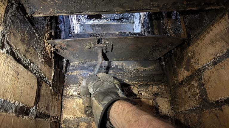 A gloved hand works the handle of a sooty throat damper inside a tan brick fireplace.