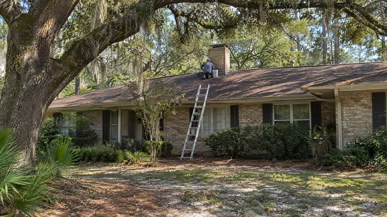 A ladder at the eave of a tan brick ranch home with a worker on the roof beside the chimney under live oaks.