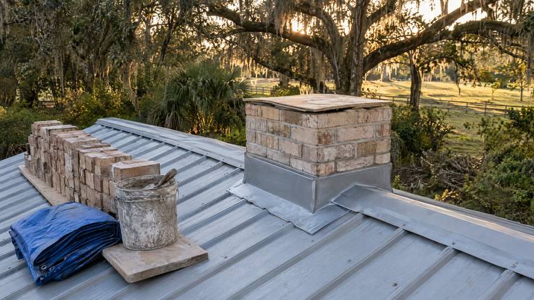 A tan brick chimney taken down to a few courses above a metal roof, its top covered with plywood, with salvaged bricks stacked on a plank beside it.