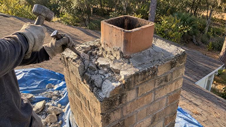 Gloved hands break up an old concrete chimney crown with a hammer and chisel, chunks piled on a tarp.