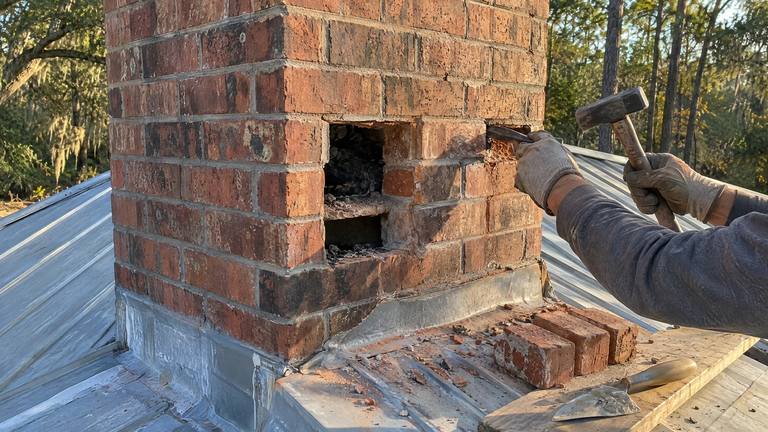 A gloved hand chisels a crumbling brick out of an orange-red chimney, new bricks waiting beside it.