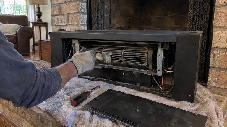 An electric fireplace insert is pulled out onto a towel while a gloved hand brushes dust from its heater fan.