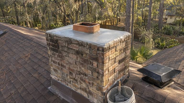 A freshly coated light gray crown on a tan brick chimney, its black cap set aside on the shingles beside a bucket.