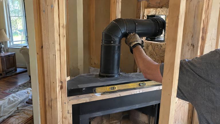 A black coaxial vent pipe elbow runs from the top of a new gas fireplace through a framed outside wall.