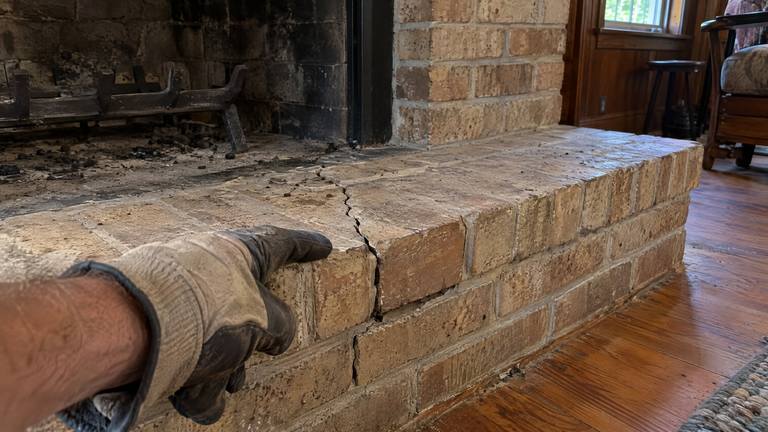 A crack runs across the bricks and joints of a raised hearth in front of a tan brick fireplace.