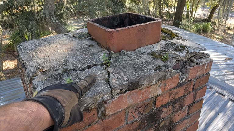 A badly cracked concrete chimney crown with small weeds growing in the cracks around a clay flue.