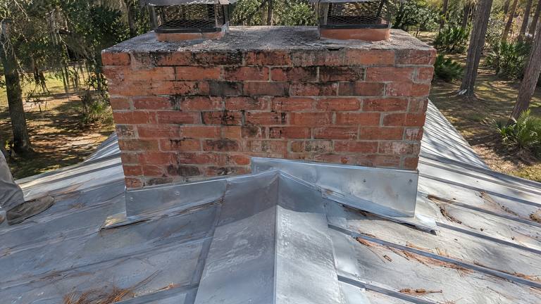 New metal flashing and a small cricket on the uphill side of a wide orange-red brick chimney with two flue caps on a metal roof.