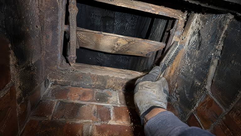 A gloved hand scrubs soot and rust off a cast-iron damper frame inside a fireplace with a wire brush.