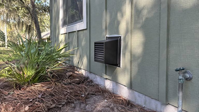 A square black direct-vent cap on the painted board-and-batten wall of a house under pine trees.
