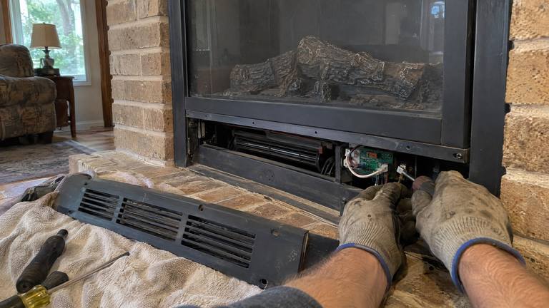 Gloved hands work at the wiring behind the lower grille of an electric fireplace insert, the grille set on a towel.