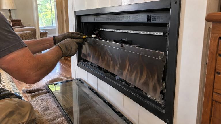An electric fireplace with its glass front off and its flame panel tilted forward while gloved hands work on a wire connector.