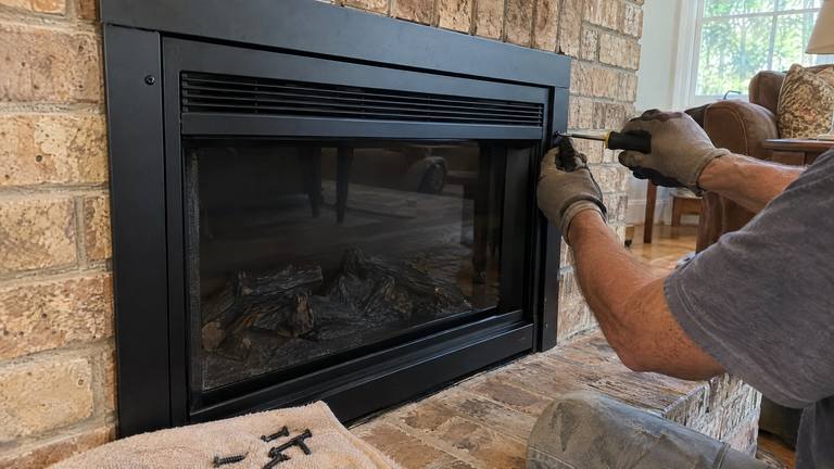 Gloved hands fasten the black trim frame of an electric insert set in a tan brick fireplace.