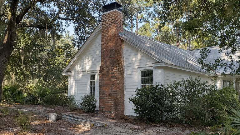 A tall red brick chimney with a black cap on the gable end of a white frame house, a ladder lying on the ground below.