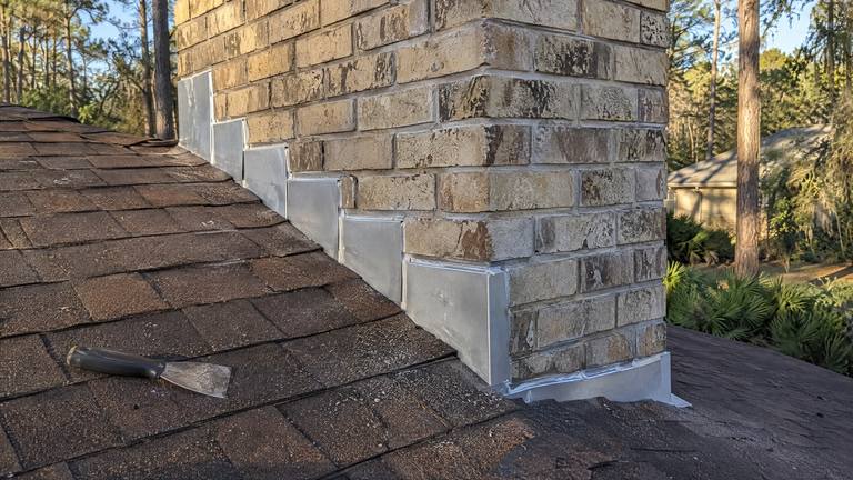 Clean new counter flashing steps down the side of a tan brick chimney over brown shingles.
