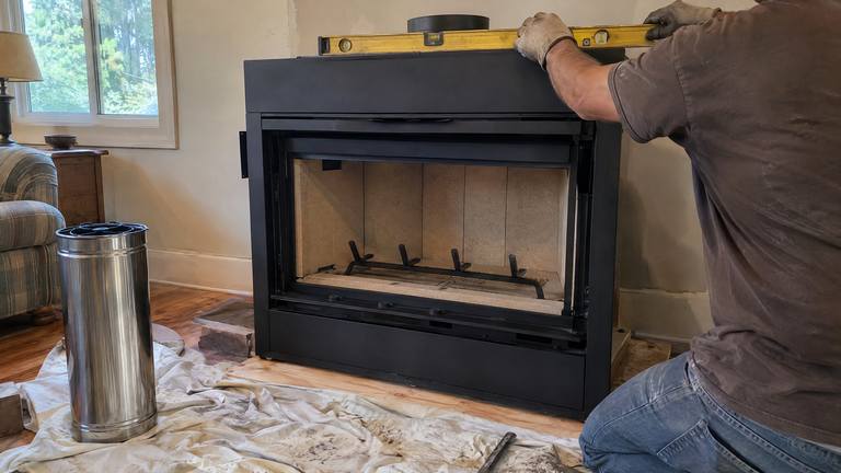 Gloved hands set a level on top of a new factory-built fireplace before the wall is framed around it.