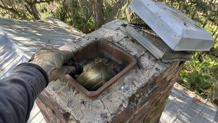 A gloved hand shines a flashlight down a square clay flue at the top of an orange-red brick chimney.