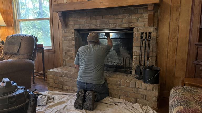 A worker kneels at a tan brick fireplace, shining a flashlight up through the open damper.