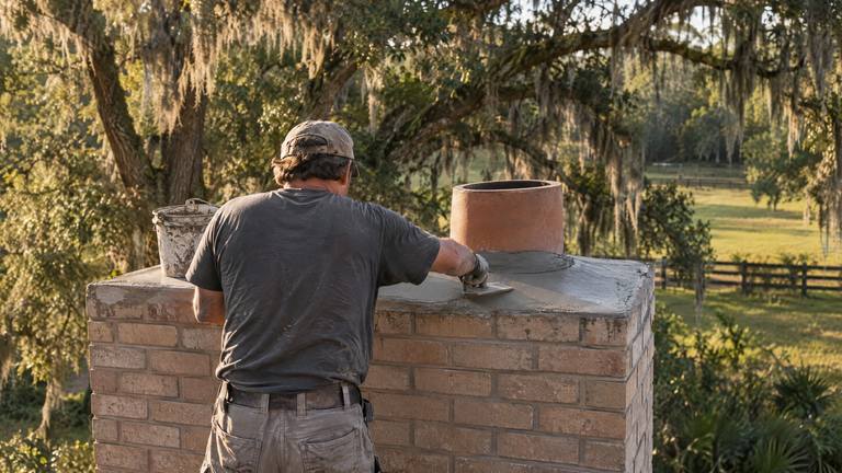 A worker trowels a new cement crown around the clay flue at the top of a tan brick chimney.