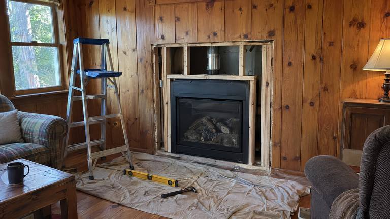 A new gas fireplace sits in a fresh framed opening in a knotty pine paneled wall, trim not yet on.