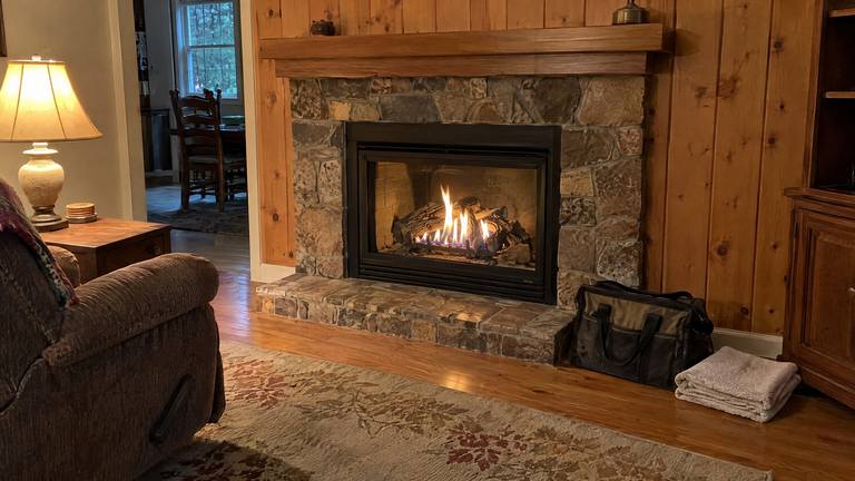 A gas fireplace with a stone surround burns evenly in a pine-paneled den, a tool bag and towel on the floor beside it.