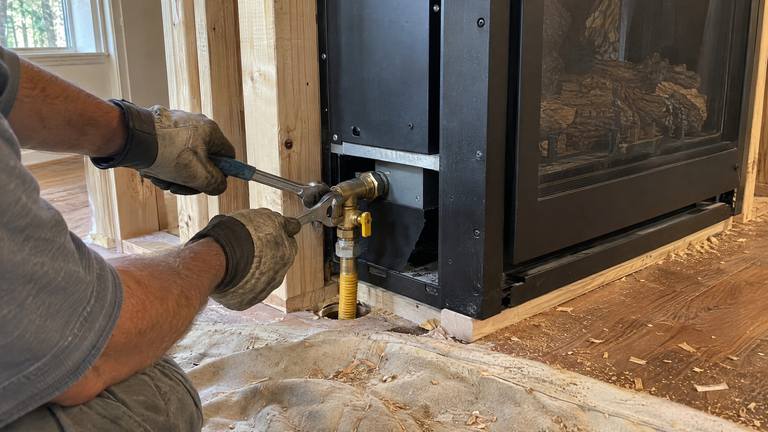 Gloved hands tighten a flexible gas line and shutoff valve on a new gas fireplace in fresh framing.