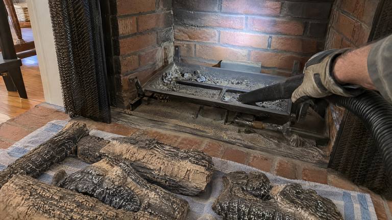 A vacuum nozzle cleans a gas log burner pan while the ceramic logs lie on a towel in front of the fireplace.