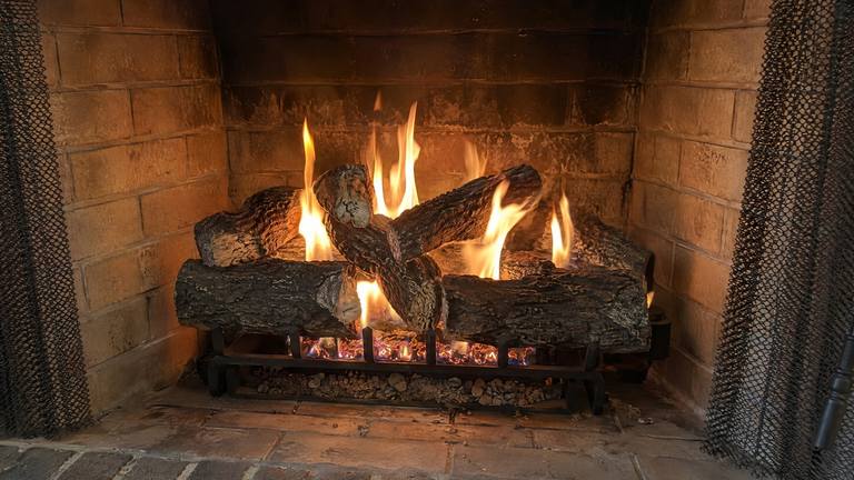 Lit gas logs on a black grate in a tan brick firebox, flames rising between the logs over glowing embers.