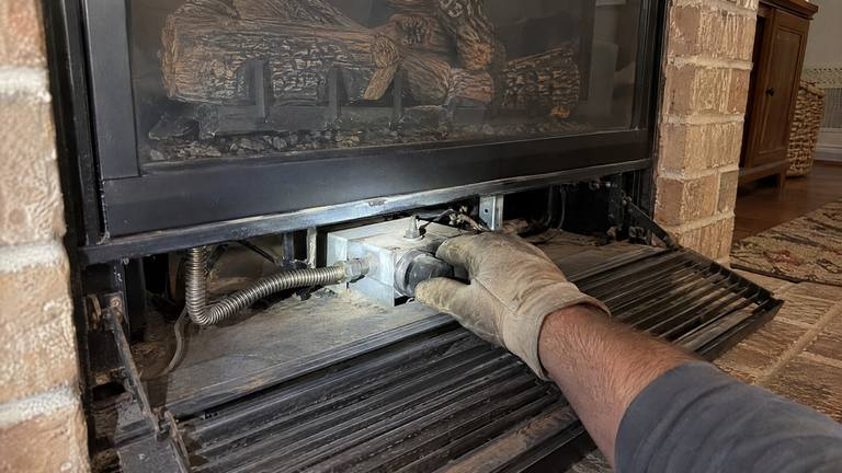 A gloved hand turns the control knob on a gas valve behind the open louver of a gas fireplace.