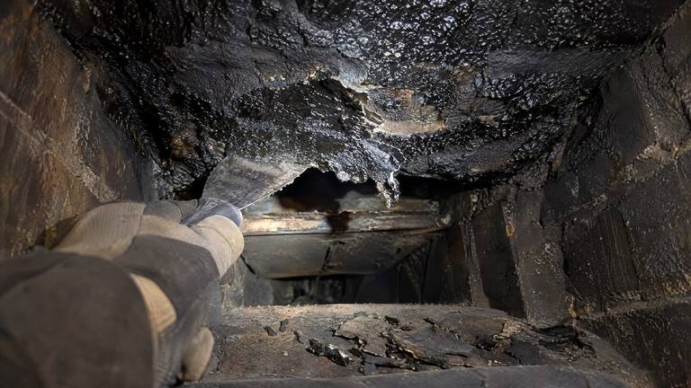 A gloved hand holds a scraper against shiny black glazed creosote coating the inside of a flue.