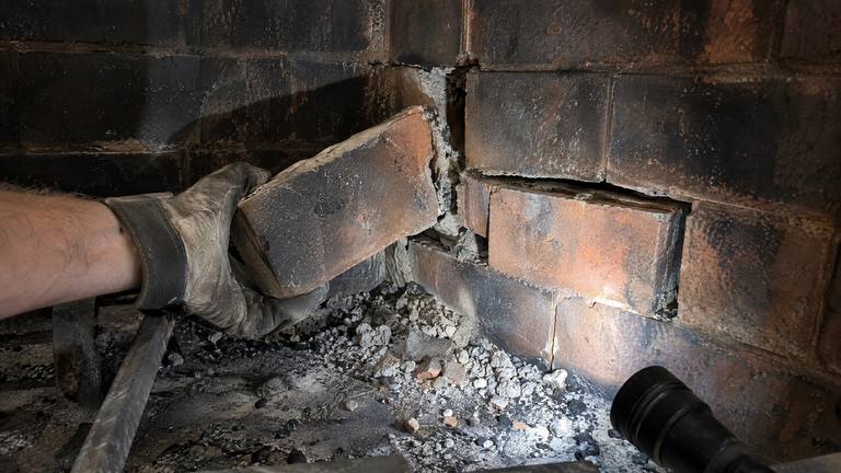 A gloved hand pulls a loose firebrick out of the sooty side wall of a firebox.