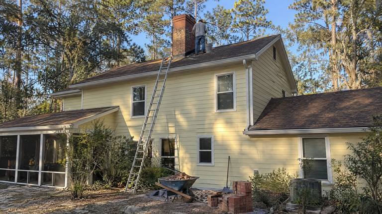 A worker stands on the roof beside the chimney of a pale yellow two-story house, with a ladder, a wheelbarrow of old bricks and new bricks below.