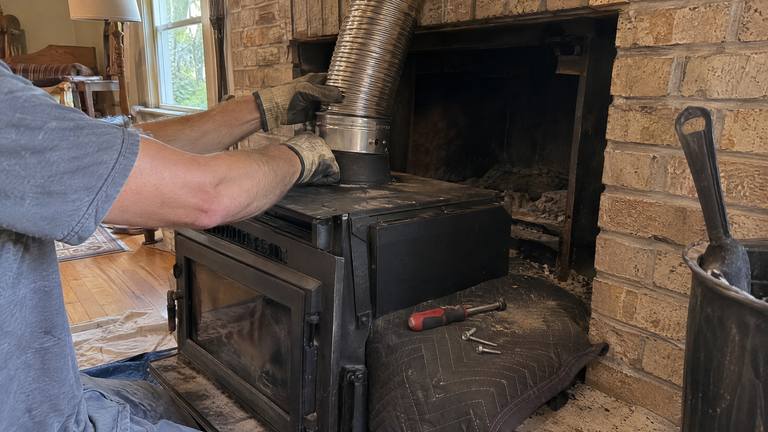 Gloved hands fit a stainless liner onto the top collar of a black wood insert pulled out onto a padded hearth.