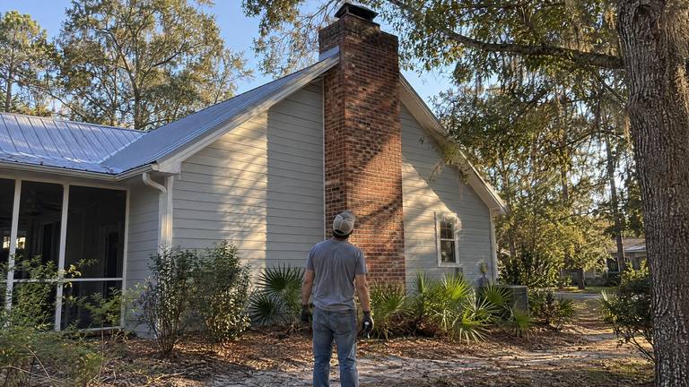 A worker stands in a sandy yard looking up along a tall exterior brick chimney on a frame house.