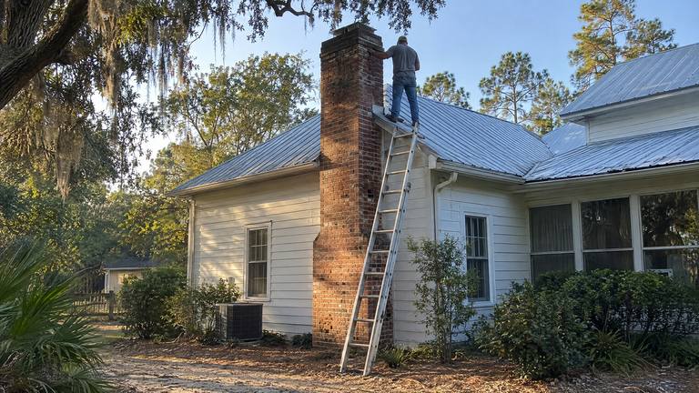 A worker stands on a metal roof beside the tall brick chimney of a white frame house, checking its top.