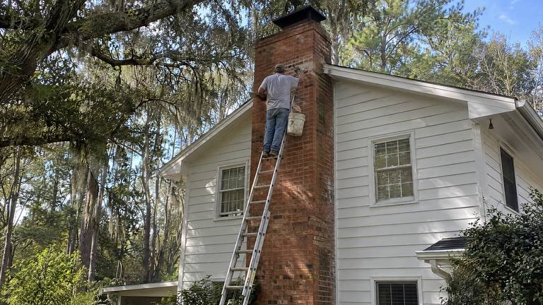 A worker on a tall ladder repairs brick halfway up the exterior chimney of a white frame house.