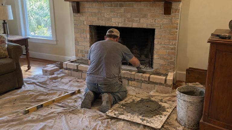 A worker lays tan bricks for a raised hearth in front of a newly installed fireplace.