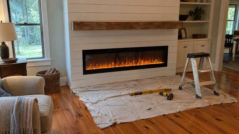 A long electric fireplace glows in a new white shiplap wall, with a drop cloth and level on the floor.