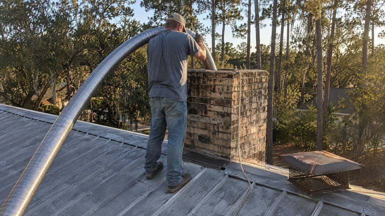 A worker on a metal roof feeds a flexible stainless liner down into a tan brick chimney.