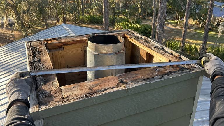 Gloved hands stretch a tape measure across the open, rotted top of a chimney chase around a metal flue pipe.