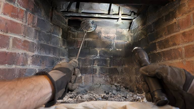 Gloved hands hold a small mirror and a flashlight inside a sooty orange-red brick firebox, angled up at the flue.