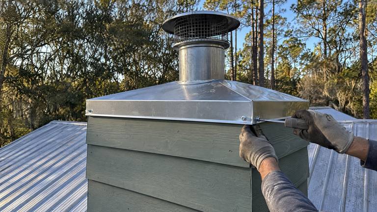 Gloved hands fasten the skirt of a new stainless chase cover on a chimney chase clad in sage green siding.