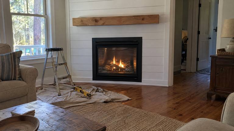 A newly installed gas fireplace burns in a white shiplap wall with a simple pine mantel.